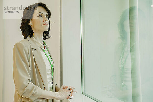 Confident businesswoman looking out through window standing with hands clasped at office