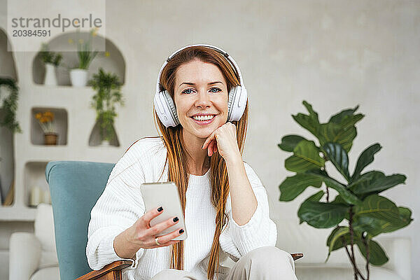 Happy woman holding smart phone listening music through headphones at home