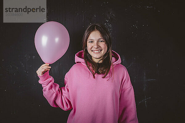 Girl in hoodie holding pink balloon in front of black background