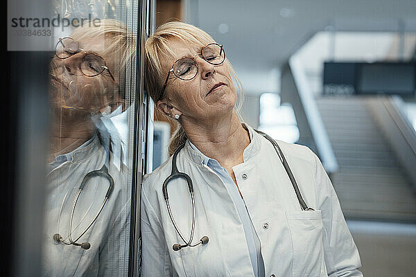 Mature doctor leaning with eyes closed on vending machine in hospital