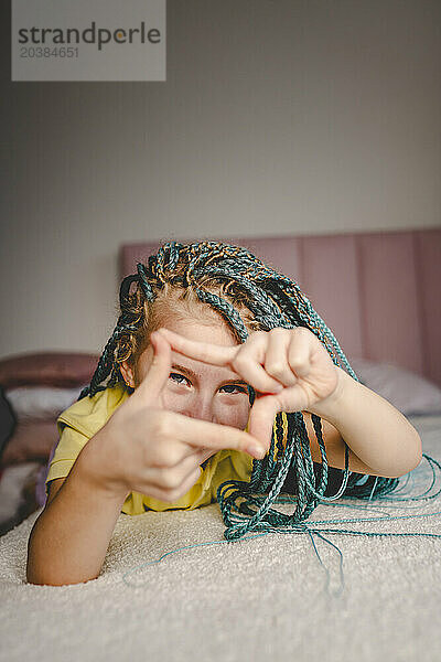 Happy girl with turquoise dyed braided hair lying on bed making finger frame at home