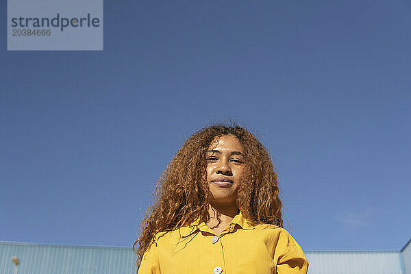 Young curly haired woman wearing yellow casuals under clear blue sky on sunny day