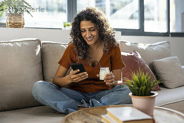 Happy woman holding yogurt and using smart phone at home