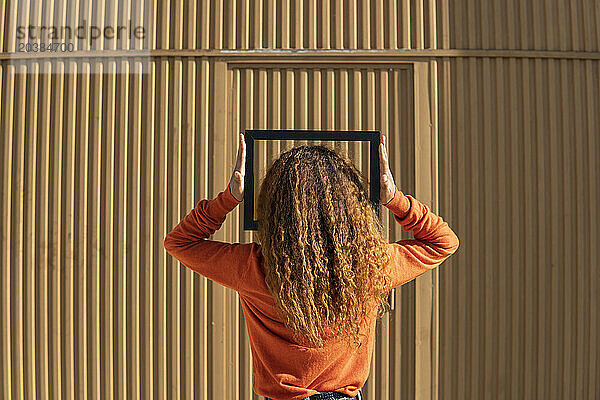 Young woman with curly hair holding picture frame standing in front of metal wall