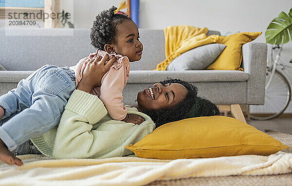 Happy young mother lying with daughter by sofa in living room