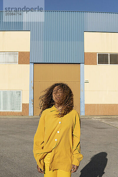 Young curly haired woman in yellow casuals with eyes closed standing on footpath