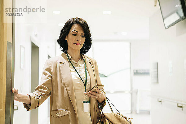 Confident mature businesswoman using mobile phone standing at illuminated office