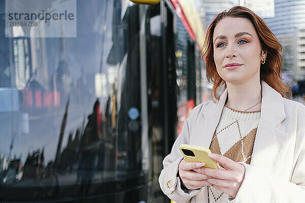 Smiling woman standing with smart phone near bus