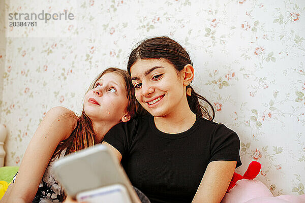 Smiling girl using smart phone near friend at home