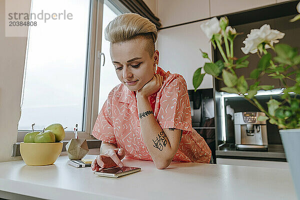 Woman leaning on elbow and scrolling smart phone in kitchen at home