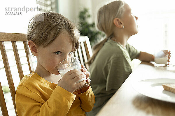 Boy drinking milk sitting next to sister at table in home