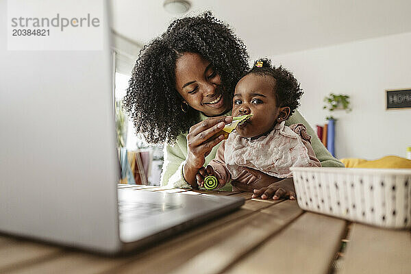 Smiling businesswoman feeding daughter with package food sitting at table