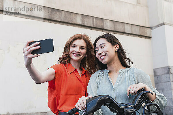 Woman taking selfie with friend through smart phone at bicycle parking station