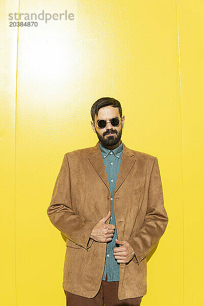 Smiling man wearing brown jacket and sunglasses near yellow wall