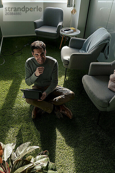 Happy businessman sitting with tablet PC on green carpet
