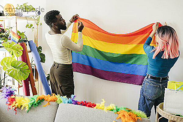 Young non-binary person sticking rainbow flag on wall with friend at home