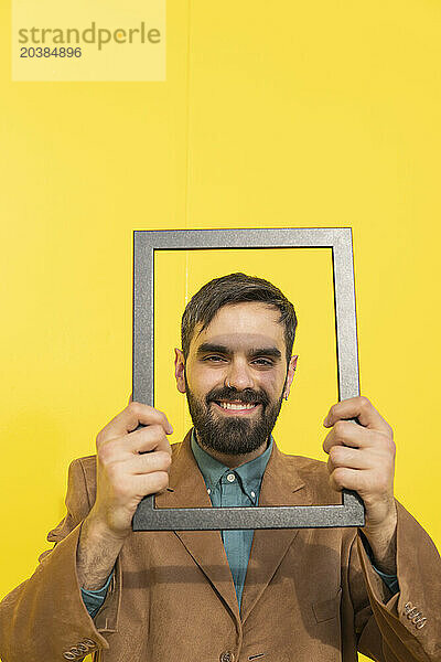 Happy man looking through picture frame against yellow background