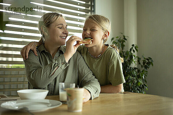 Smiling woman feeding sandwich to daughter at home