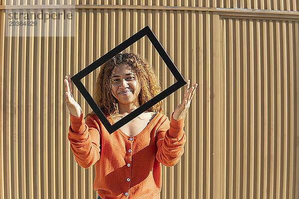 Smiling young woman holding picture frame standing in front of metal wall