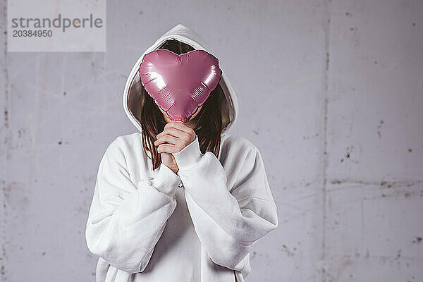 Girl covering face with heart shaped balloon in front of gray cement background