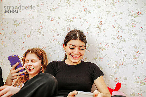 Smiling friends using smart phone near wall at home