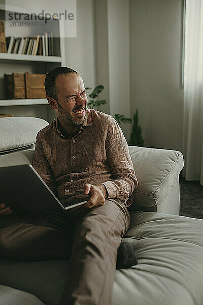 Cheerful freelancer sitting with laptop on sofa at home office