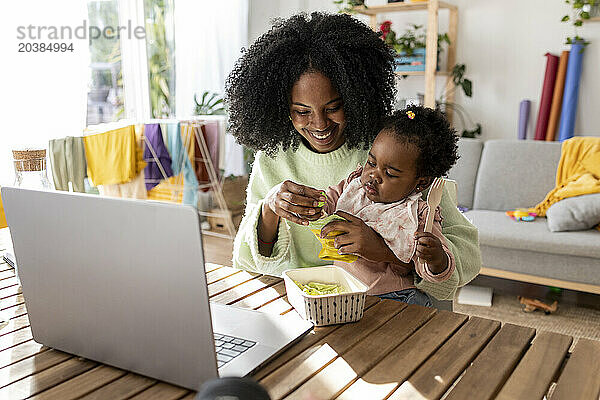 Smiling businesswoman assisting daughter eating package food sitting at table