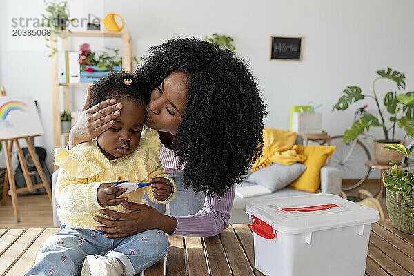 Young mother kissing daughter playing with thermometer by basket on table at home