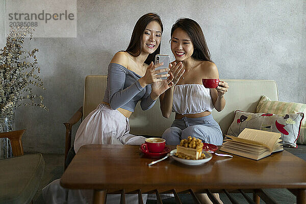Happy young friends sharing smart phone sitting on sofa at restaurant
