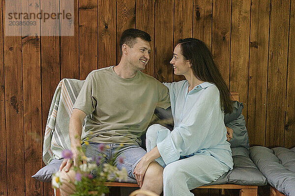 Romantic couple looking at each other sitting on sofa in front of wooden wall at home