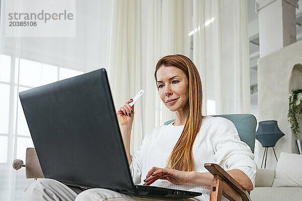 Woman shopping online through laptop sitting in chair at home
