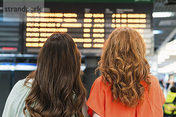 Young redhead woman looking at arrival departure board with friend at airport