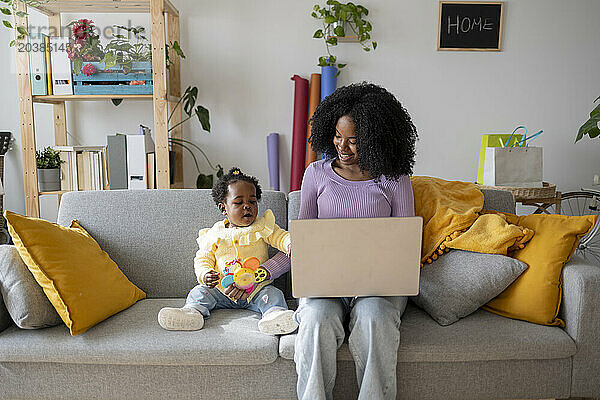 Smiling young mother using laptop working at home playing with baby girl on sofa in living room