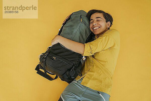 Happy woman embracing backpack against yellow background