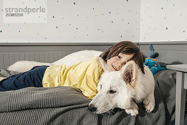 Smiling boy lying with white Swiss shepherd dog on bed at home