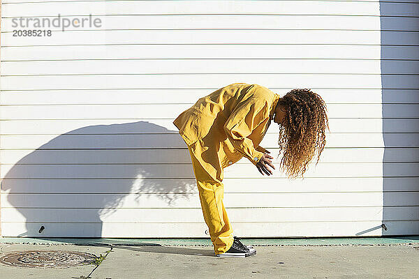 Young woman in yellow casuals with curly hair bending forward by white shutter