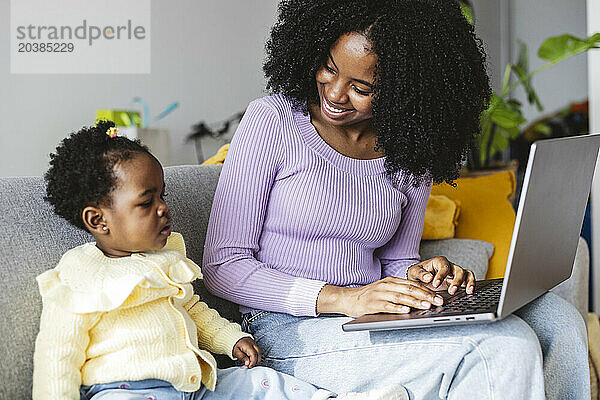 Smiling businesswoman sitting on sofa with laptop and daughter working at home