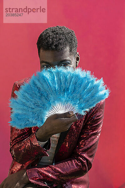 Young non-binary person holding blue hand fan against pink background