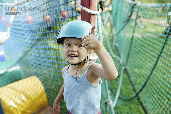 Cute girl wearing helmet and showing thumbs up gesture at rope park