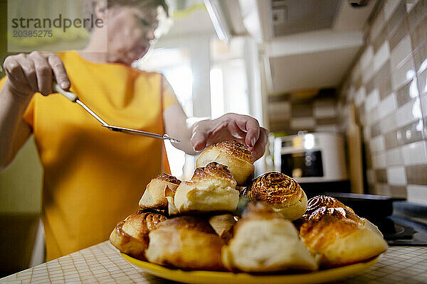 Senior woman putting sweet buns on plate in kitchen at home