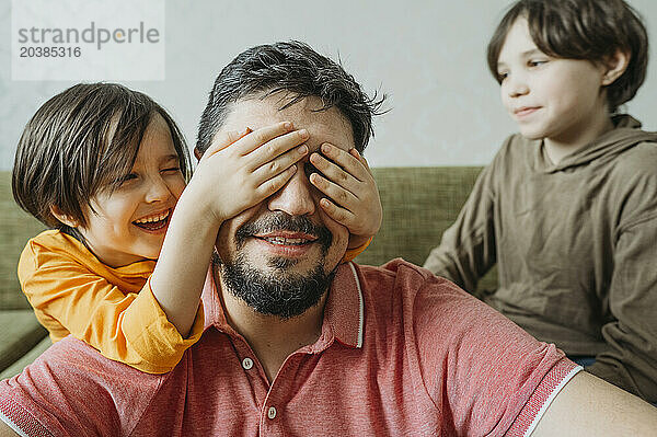 Playful boy covering father's eyes near brother at home