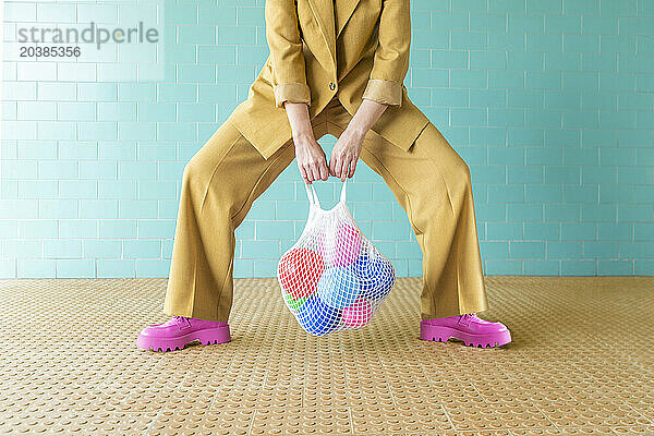 Woman holding mesh bag with colorful balloons near turquoise wall