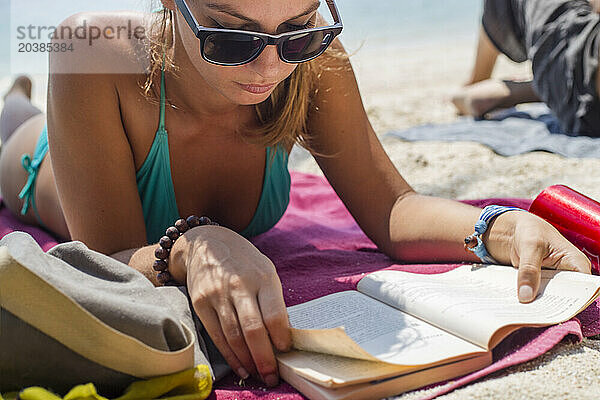 Woman wearing sunglasses and reading book lying down at beach