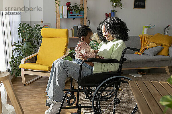 Baby girl sitting on smiling mother with disability in wheelchair at home