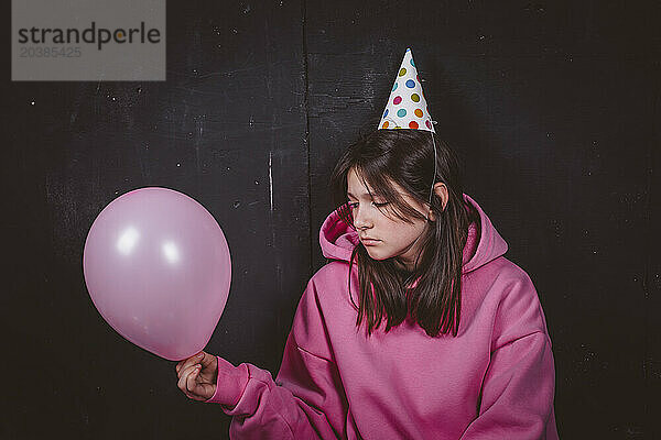 Sad birthday girl in party hat looking at pink balloon in front of black background