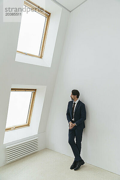 Businessman leaning on white wall looking out through window at office