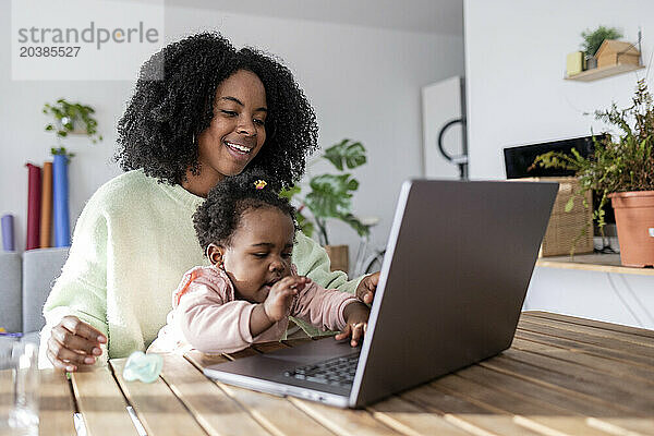 Happy working mother looking at daughter playing with laptop in home