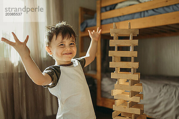 Confident boy with arms raised near wooden block tower at home