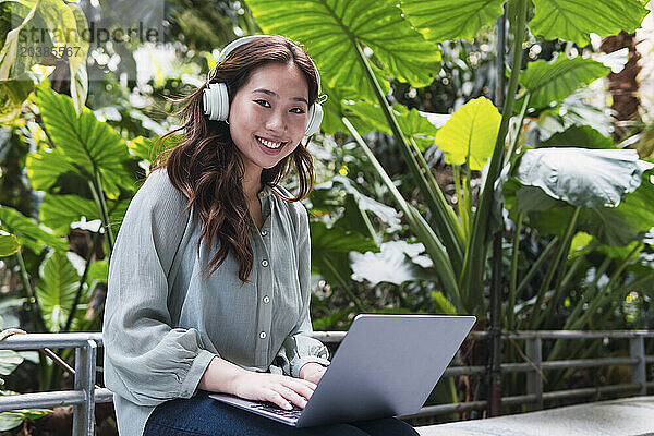 Smiling woman with laptop and wireless headphones sitting on bench