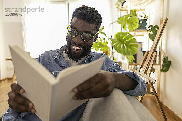 Smiling non-binary person wearing eyeglasses and reading book at home